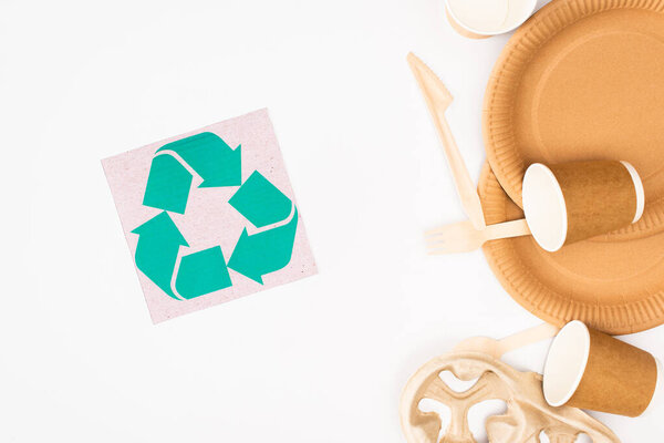 Top view of card with recycle sign and disposable tableware and wooden cutlery on white background, ecology concept