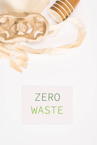 Top view of bag, paper cups and wooden cutlery near card with zero waste lettering on white background, ecology concept