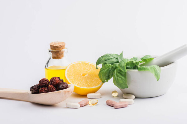 green leaves in mortar, berries and lemon near pills on white background, naturopathy concept