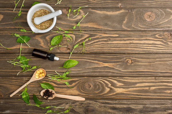 top view of pills in spoons, mortar, green herbs and bottle on wooden surface, naturopathy concept