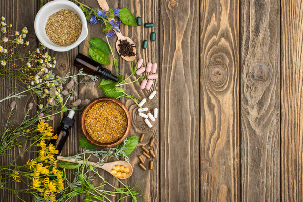 top view of pills in spoons, green herbs and wildflowers on wooden surface, naturopathy concept