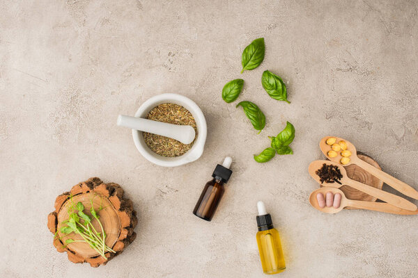 top view of herbs, green leaves, mortar with pestle, bottles and pills in wooden spoons on concrete background, naturopathy concept