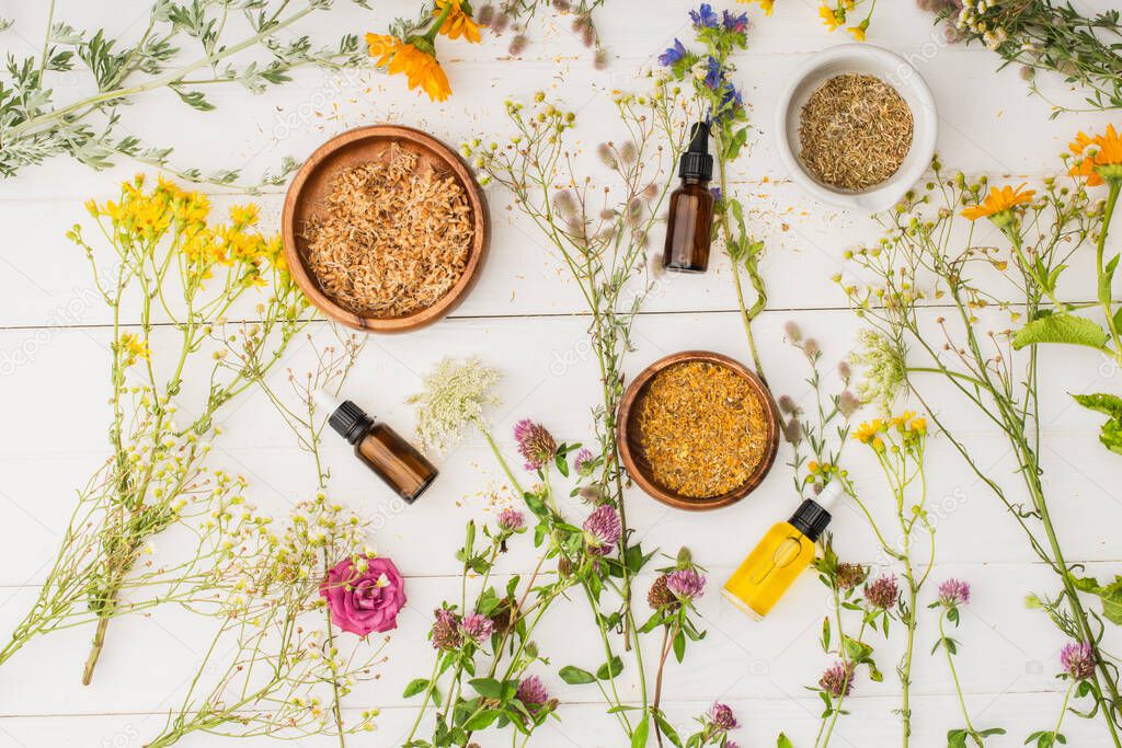 top view of herbs in bowls near flowers and bottles on white wooden background, naturopathy concept