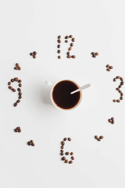 top view of clock made of coffee beans around cup on white surface