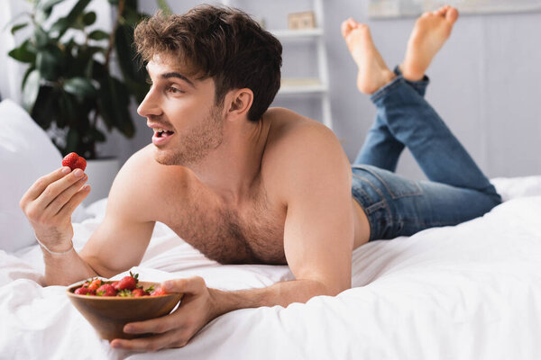 selective focus of shirtless and barefoot man lying on bed and holding bowl with ripe strawberries 