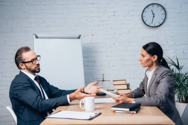 Gözlüklü bir işadamı resmi olarak iş kadınının yanında el kol hareketi yapıyor. Bardak, not defteri ve not panosunun yanında takıyor.