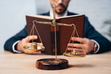 cropped view of bearded lawyer reading book near golden scales on table clipart