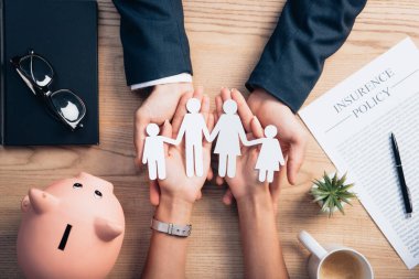 top view of lawyer and woman holding paper cut family near cup of coffee, insurance policy agreement, piggy bank and plant clipart