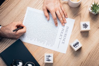 cropped view of lawyer signing insurance policy agreement near plant, glasses, notebook and wooden cubes with family, car and house clipart