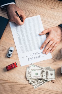 partial view of lawyer signing insurance policy agreement near dollars and toy cars clipart