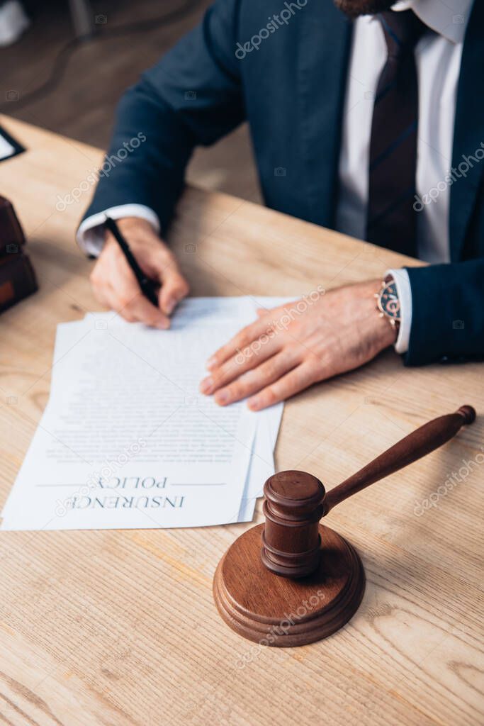 selective focus of judge signing papers with insurance policy lettering near gavel on table