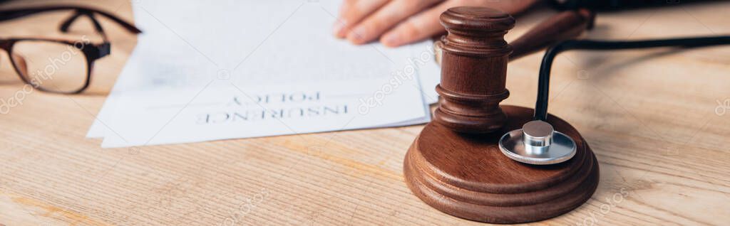 panoramic crop of judge near papers with insurance policy lettering and gavel on table