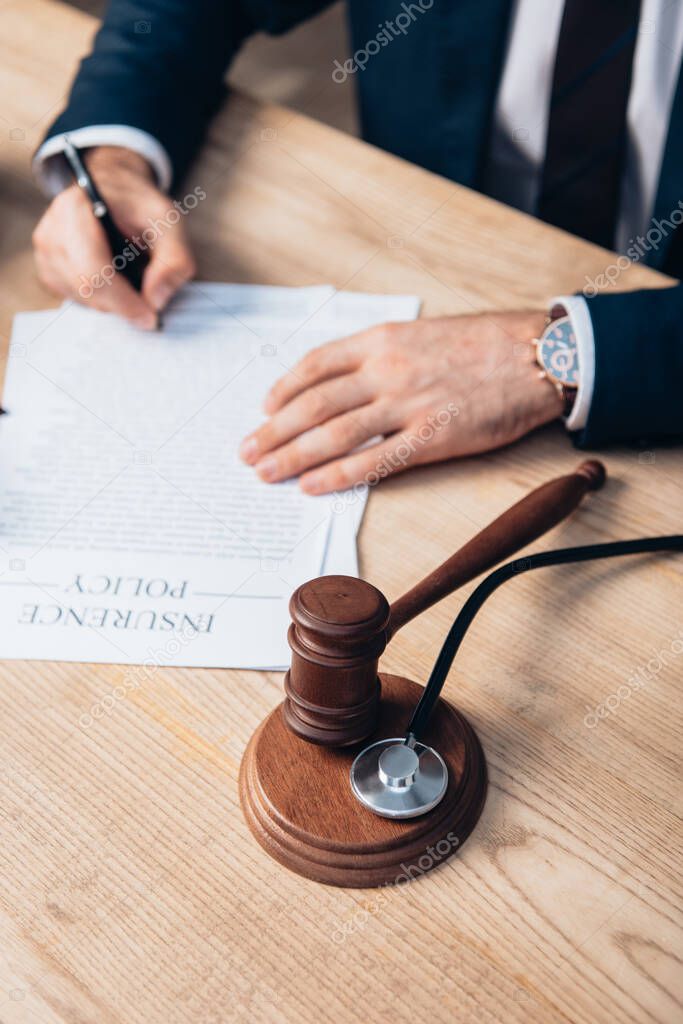 partial view of judge signing papers with insurance policy lettering near gavel and stethoscope on table