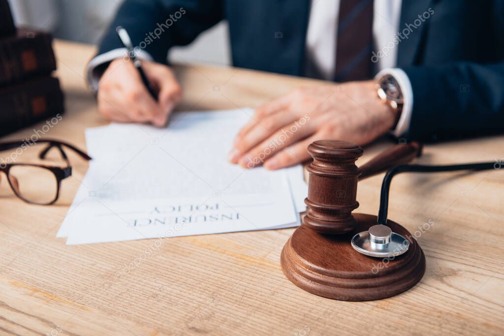 cropped view of judge signing papers with insurance policy lettering near gavel and stethoscope on table