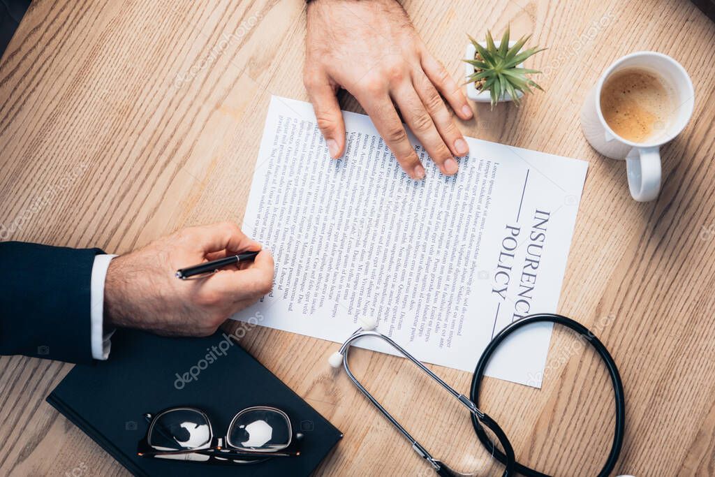 cropped view of lawyer signing insurance policy agreement near plant, glasses, notebook and stethoscope on desk