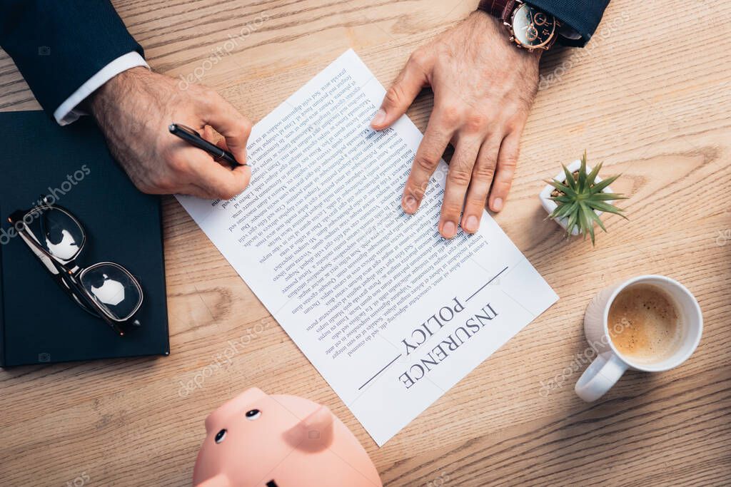 top view of lawyer signing insurance policy agreement near plant, glasses, notebook and piggy bank on desk