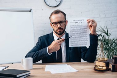 selective focus of lawyer in glasses holding pen and contract with insurance policy lettering while looking at camera  clipart