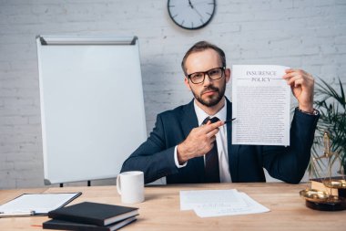 selective focus of bearded lawyer in glasses holding pen and contract with insurance policy lettering in office clipart