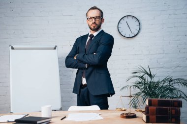 bearded lawyer in glasses standing with crossed arms near table in office clipart