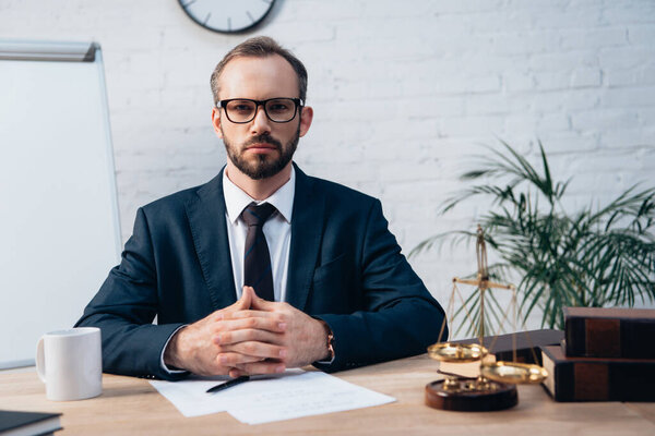 bearded lawyer in glasses looking at camera and sitting with clenched hands at desk