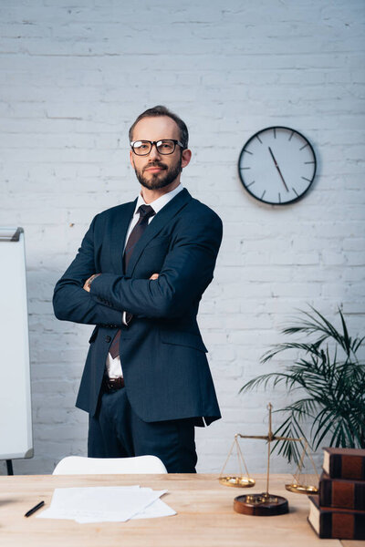 lawyer in glasses standing with crossed arms near table in office
