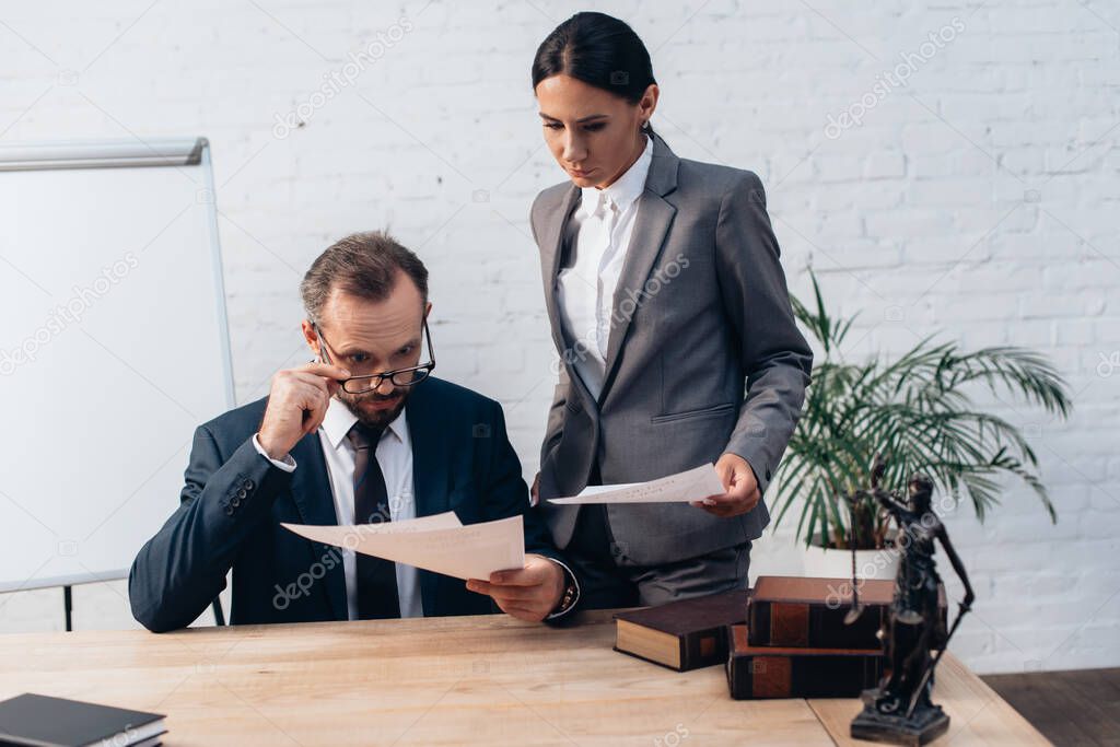 bearded lawyer in glasses looking at documents near coworker in office