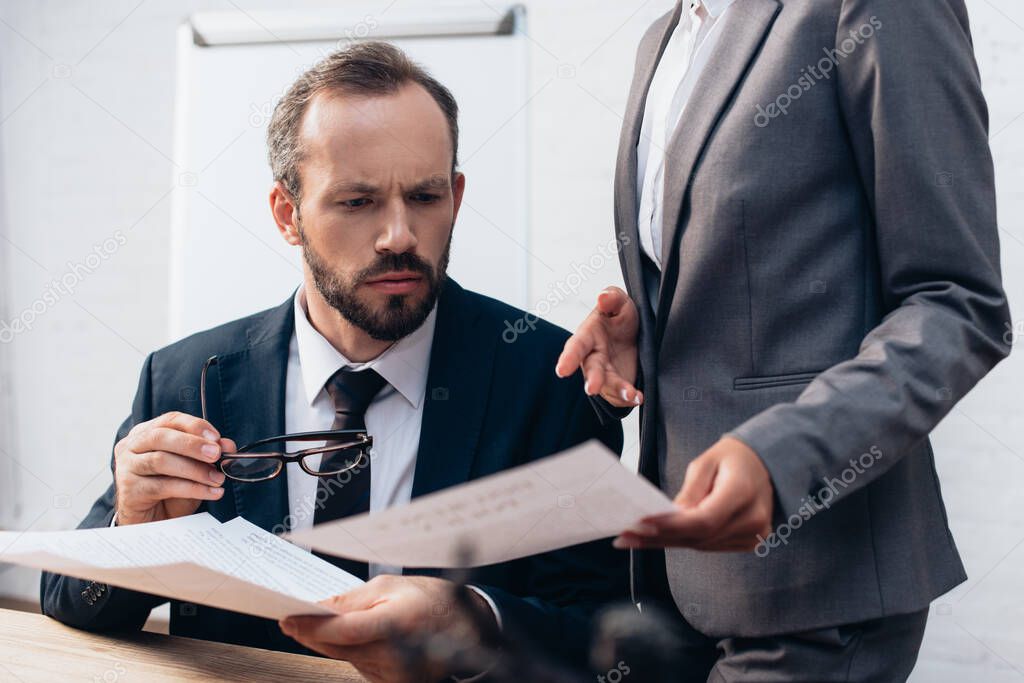 selective focus of bearded lawyer in glasses looking at documents while coworker pointing with hand in office