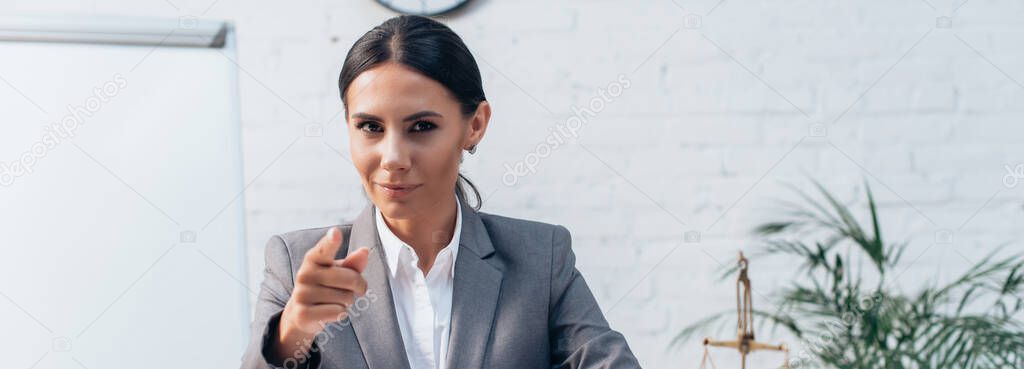 panoramic crop of brunette lawyer in formal wear looking at camera and pointing with finger in office