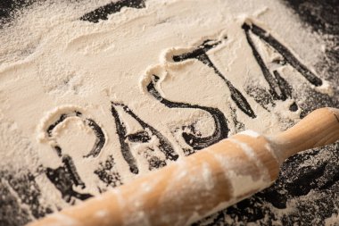 pasta lettering written on flour near rolling pin on black background clipart