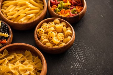 Tahta kaselerde siyah arka planda çeşitli çiğ İtalyan makarnalarının seçici odağı.