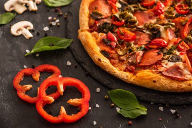 Siyah arka planda sebzelerin yanında salamlı lezzetli İtalyan pizzasının yakın görüntüsü.