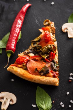 Lezzetli İtalyan pizza diliminin üst görüntüsü. Siyah arka planda sebzelerin yanında salam var.