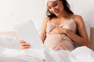 Seçici odak noktası ipek gecelikli hamile kadın dijital tablet tutuyor ve yatak odasında karnına dokunuyor.