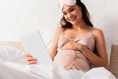 Seçici odak noktası ipek gecelikli hamile kadın dijital tablet tutuyor ve yatak odasında karnına dokunuyor.