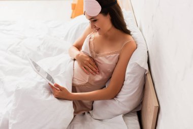 ipek gecelikli hamile kadın ve göz maskesi takan dijital tablet tutan ve yatak odasında karnına dokunan.