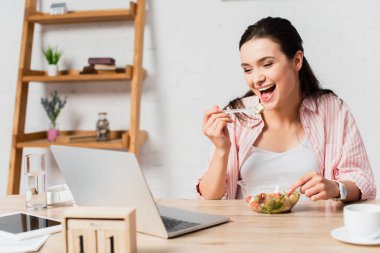 Esmer kadın salatanın yanında dizüstü bilgisayara, tablete ve masaya bakıyor. 