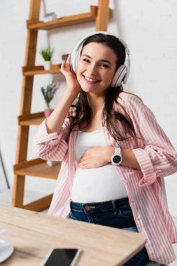 Kablosuz kulaklıkla akıllı telefonun yanında boş ekranlı müzik dinleyen hamile kadının seçici odağı. 