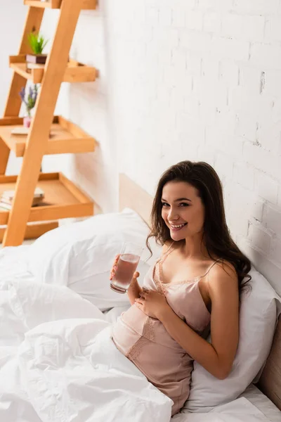 年轻的孕妇 手里拿着一杯水 在卧室里摸着肚子 — 图库照片