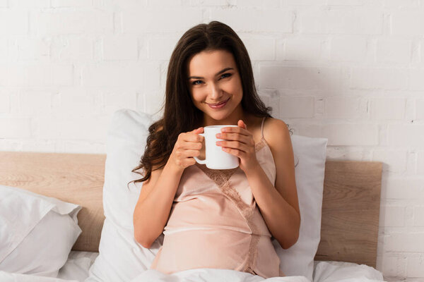 young and pregnant woman in silk nightie holding cup of tea 