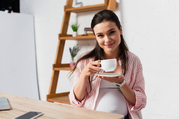 selective focus of pregnant woman looking at camera while holding cup of tea