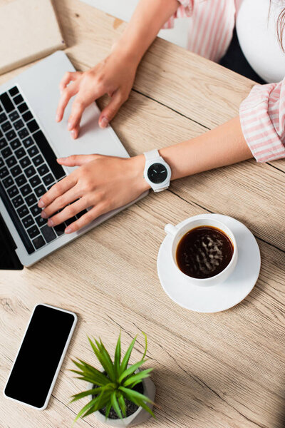 top view of freelancer using laptop near cup of coffee, plant and smartphone with blank screen 