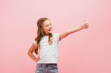 girl in white t-shirt with hand on hip looking away while showing thumb up isolated on pink clipart
