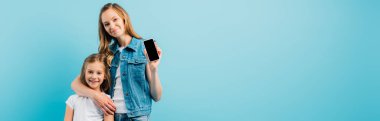 Kotlu yelek giyen annenin yatay görüntüsü mavi renkte izole edilmiş kızı kucaklarken boş ekranlı akıllı telefonu gösteriyor.