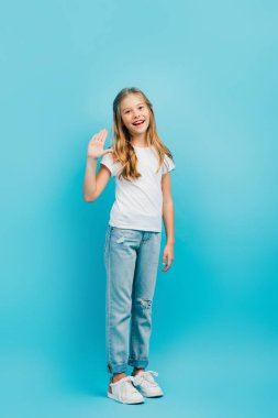 Beyaz tişörtlü ve kot pantolonlu bir kızın mavi kameraya bakarken el sallaması. 