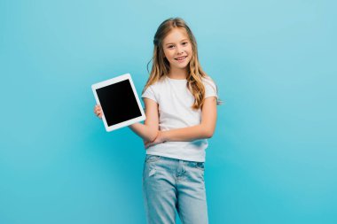 Beyaz tişörtlü ve kot pantolonlu çocuk mavi üzerine izole edilmiş kameraya bakarken dijital tablet gösteriyor