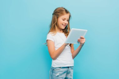 Dijital tablet kullanan beyaz tişörtlü ve kot pantolonlu çocuk mavi üzerinde tek başına duruyor