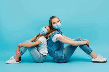 Kot elbiseli ve tıbbi maskeli genç bir kadın ve kız sırt sırta mavinin üzerinde oturuyor.