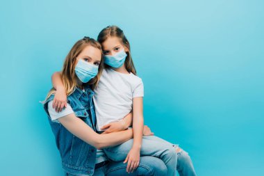 Beyaz tişörtlü çocuk, anne kucağında kot yelekli, maske takıyor ve mavi kameraya bakıyor.