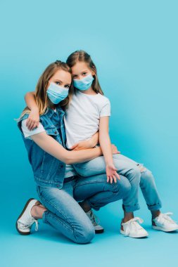 Beyaz tişörtlü bir kız. Kucağında kot giysiler içinde annesinin kucağında oturuyor. Maske takıyor ve mavi kameraya bakıyor.