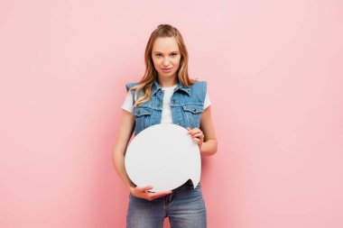 Kot yelekli genç bir kadın düşünce baloncuğu tutuyor ve pembe üzerine izole edilmiş kameraya bakıyor.